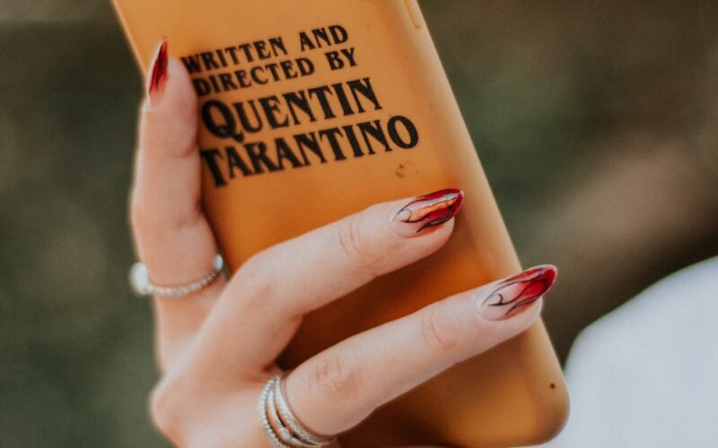 Estilo cinematográfico Tarantino — Elegant hand holding a smartphone with distinctive director-inspired phone case, showcasing nail art and rings.