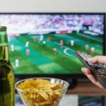 assistir jogos de futebol ao vivo — A person holding a remote while watching soccer on TV with beer and snacks on the table.