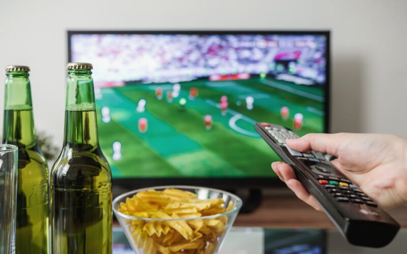 assistir jogos de futebol ao vivo — A person holding a remote while watching soccer on TV with beer and snacks on the table.
