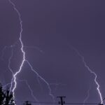 chuva forte e temporais — A powerful lightning strike illuminates the night sky over a rural landscape with silhouettes of houses and trees.