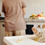 histórias de superação — Cute toddler sits in a high chair enjoying a meal, while parents cook in the background of a modern kitchen.