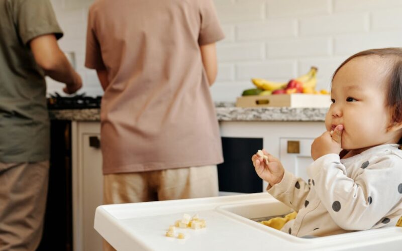 histórias de superação — Cute toddler sits in a high chair enjoying a meal, while parents cook in the background of a modern kitchen.