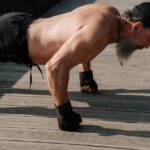 exercício físico na terceira idade — Senior man with a beard exercising outdoors, performing push-ups on a wooden deck.