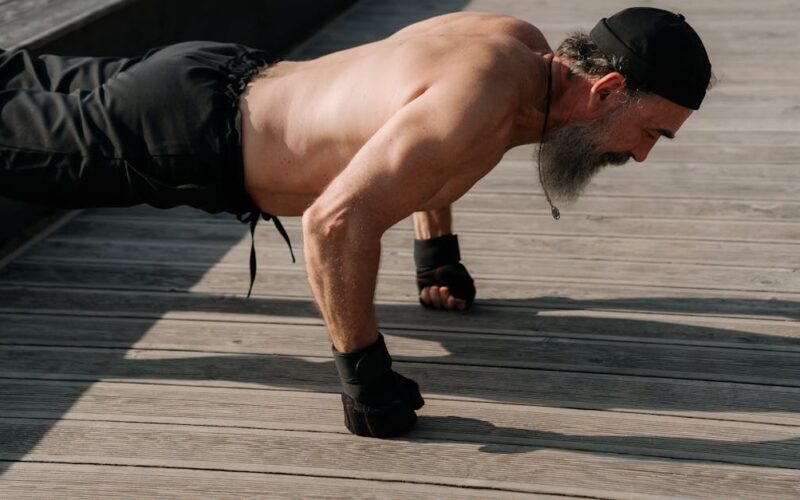 exercício físico na terceira idade — Senior man with a beard exercising outdoors, performing push-ups on a wooden deck.