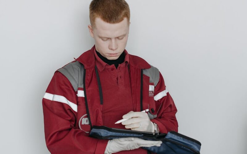 documentação essencial para emergências — A male paramedic in red uniform writing notes while sitting indoors, focused and professional.