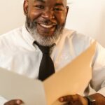 organização documentos terceira idade — Senior businessman with beard sitting at desk, smiling while holding a folder in a modern office setting.