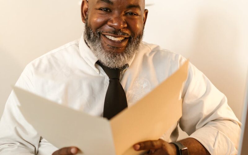organização documentos terceira idade — Senior businessman with beard sitting at desk, smiling while holding a folder in a modern office setting.