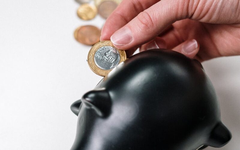 proteção investimentos previdenciários — A hand putting a coin into a black piggy bank with scattered coins on a white background.