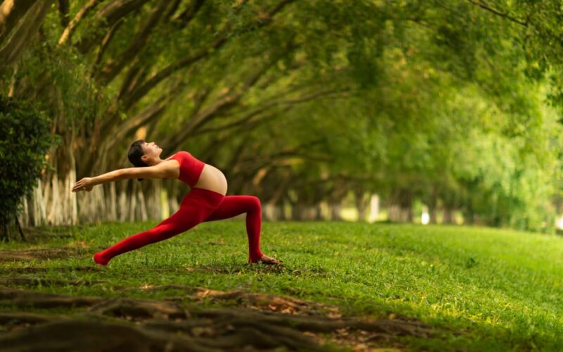 gravidez ativa segura — Pregnant woman in red practicing yoga outdoors in a lush green park, striking a pose.