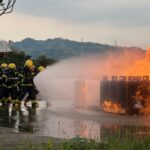 investimentos segurança civil bombeiros — Team of firefighters using hoses to extinguish a controlled outdoor blaze during training.