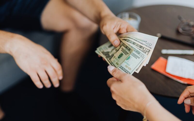quinto dia útil — Close-up of hands exchanging US dollar bills, symbolizing a financial transaction or payment.