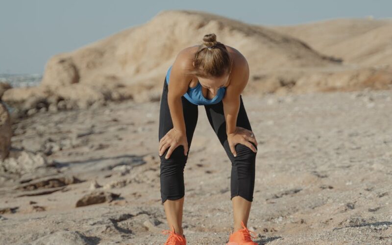 desempenho atletas copa do mundo — A tired woman in athletic wear taking a break on a sandy beach under a clear sky.