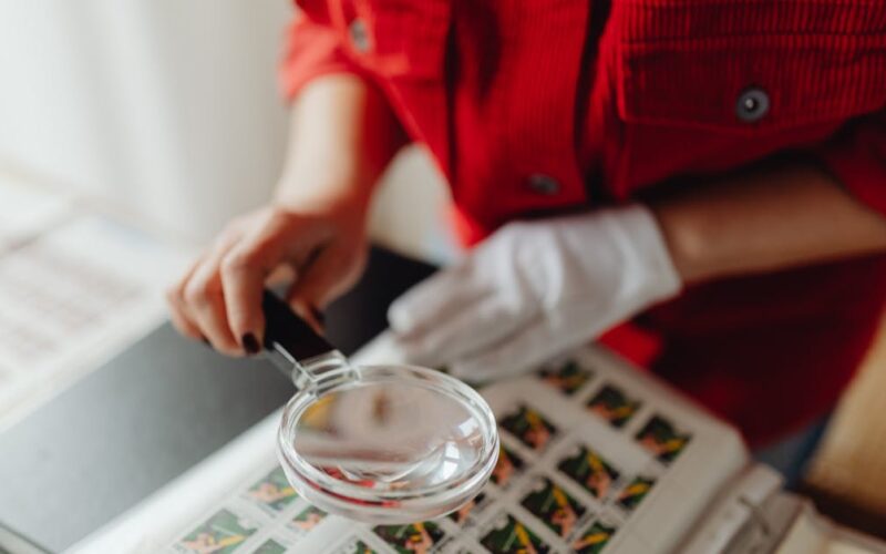 pirataria e fraudes financeiras — A person closely examines postage stamps with a magnifying glass, revealing intricate details.