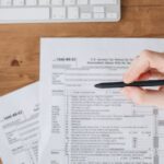 declarar ganhos apostas IR 2026 — Close-up of a hand holding a pen over US tax forms on a wooden table.