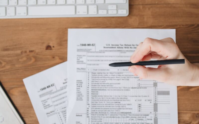 declarar ganhos apostas IR 2026 — Close-up of a hand holding a pen over US tax forms on a wooden table.