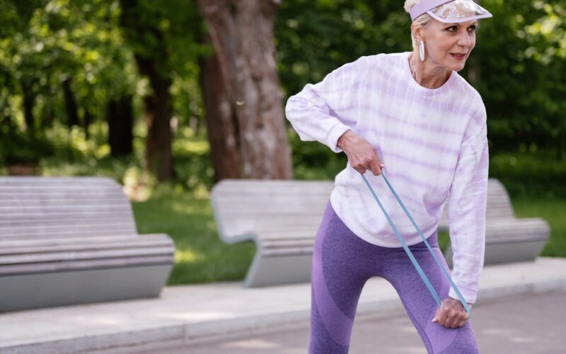 saúde mental melhor idade — Senior woman performing outdoor exercise with a resistance band in a park setting.