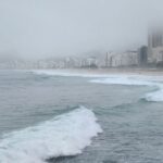 frio rigoroso no Sul do Brasil — A foggy view of the ocean with city skyline in the mist, rocky shoreline front.