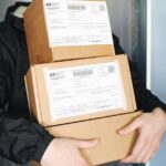 prejuízo dos Correios — Courier carrying stacked cardboard packages for delivery service indoors.