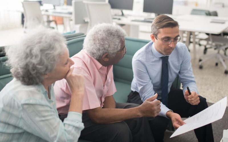 planejamento financeiro aposentadoria — Business professional consults elderly clients in an office setting. Collaborative discussion, paperwork visible.
