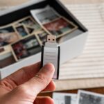 planejamento documentação familiar — Hand holding a USB drive with a box of vintage printed photos on a table, symbolizing digital preservation of memories.