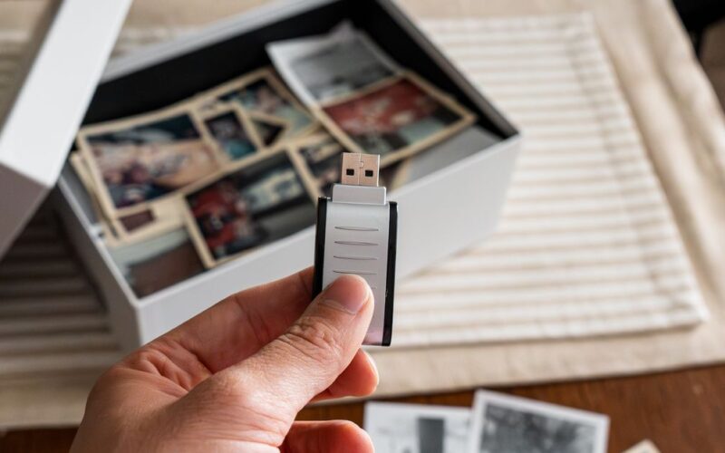 planejamento documentação familiar — Hand holding a USB drive with a box of vintage printed photos on a table, symbolizing digital preservation of memories.
