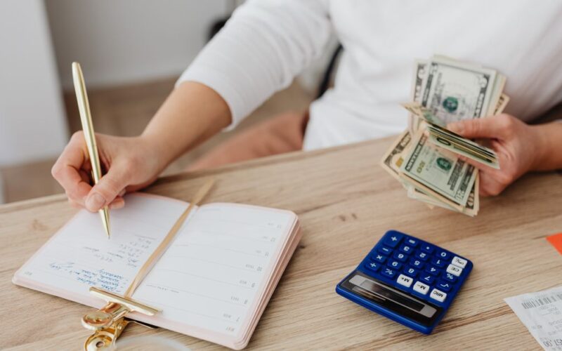 portabilidade crédito consignado — Adult holding cash and writing in planner while using a calculator at home.